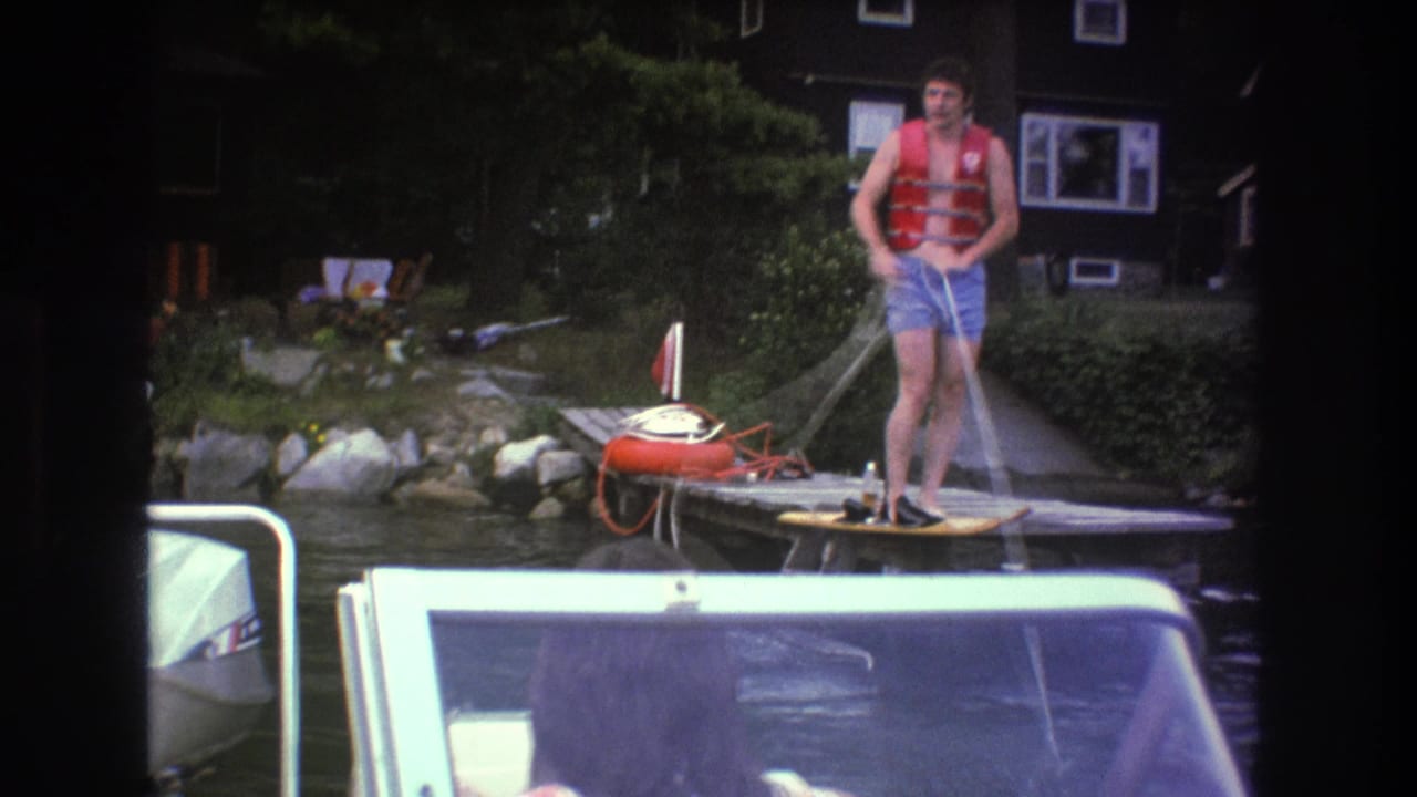 The man remains on the dock in a red life jacket, holding the water-ski tow rope beside a shoreline with shrubs, rocks, and a dark lakeside house.