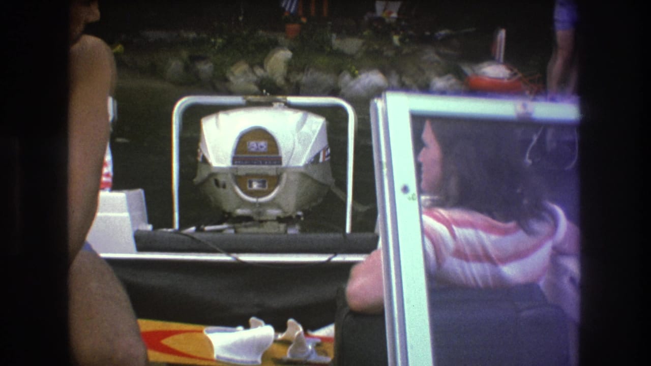 A close home-movie view from the boat shows the outboard motor, windshield frame, and partially visible adults while the dock and shoreline sit beyond the frame.