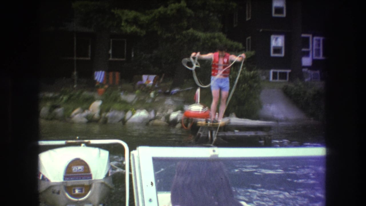 The water-skiing setup continues as the man in a red life jacket gathers the tow rope on the wooden dock, with the motorboat foreground and lakeside house behind him.