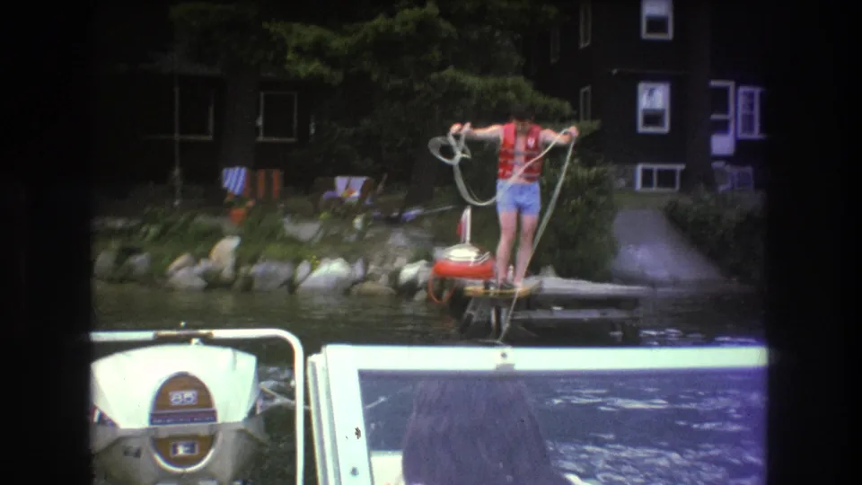 The water-skiing setup continues as the man in a red life jacket gathers the tow rope on the wooden dock, with the motorboat foreground and lakeside house behind him.