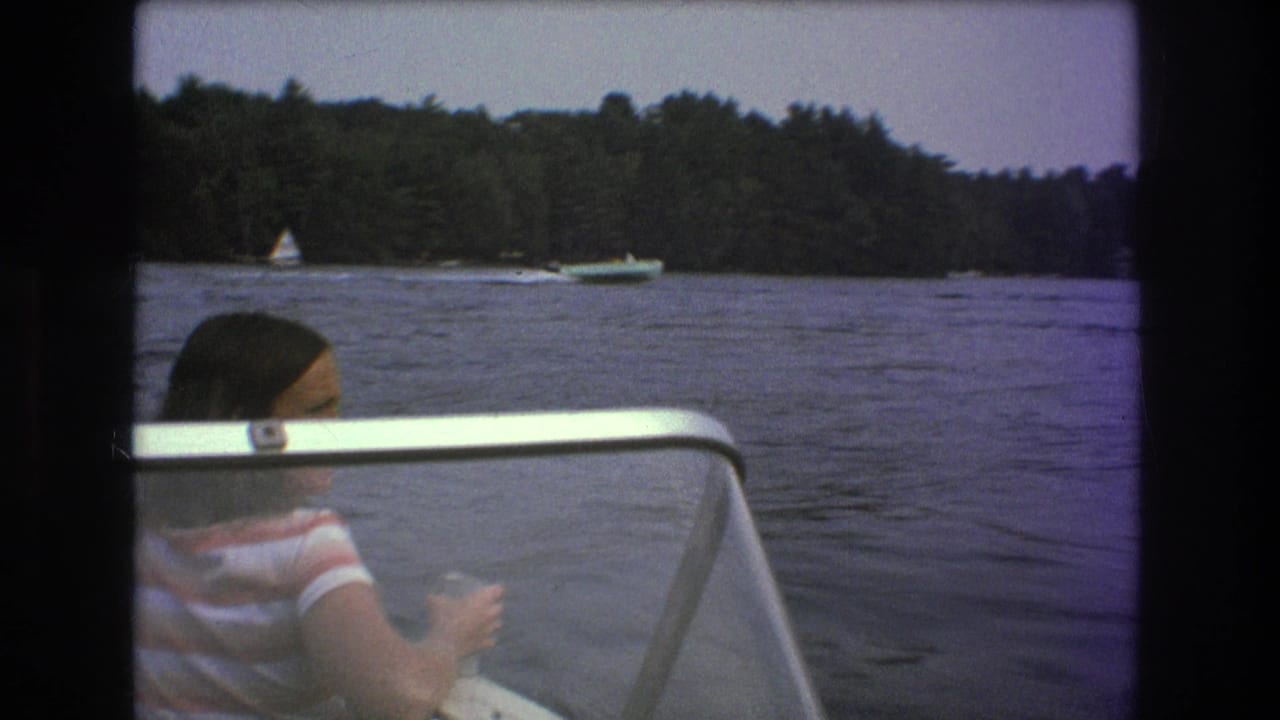 A young woman in a striped shirt sits in a small motorboat on a wooded lake in archival 8mm footage identified as Boston, United States, 1975.