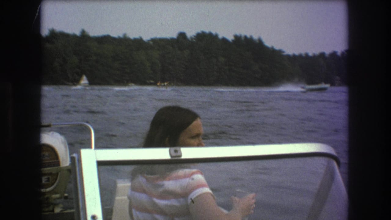 The passenger remains seated in the boat while distant sail and motorboats move across a lake near a wooded shoreline, consistent with the supplied 1975 Boston-area metadata.