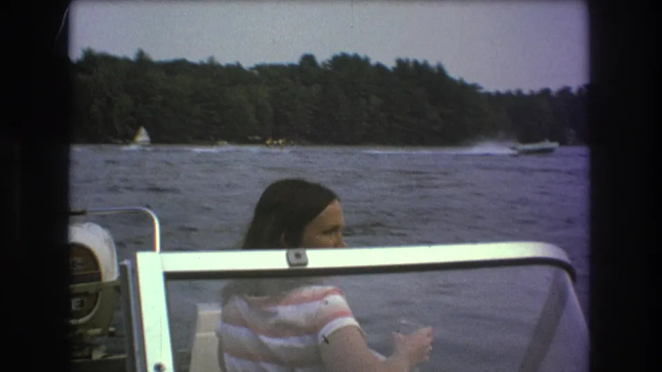 The passenger remains seated in the boat while distant sail and motorboats move across a lake near a wooded shoreline, consistent with the supplied 1975 Boston-area metadata.