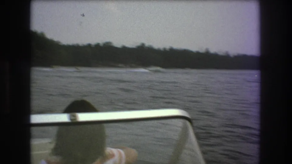 A handheld 8mm frame looks past the boat windshield and the passenger’s head toward open water, shoreline trees, and a distant motorboat.