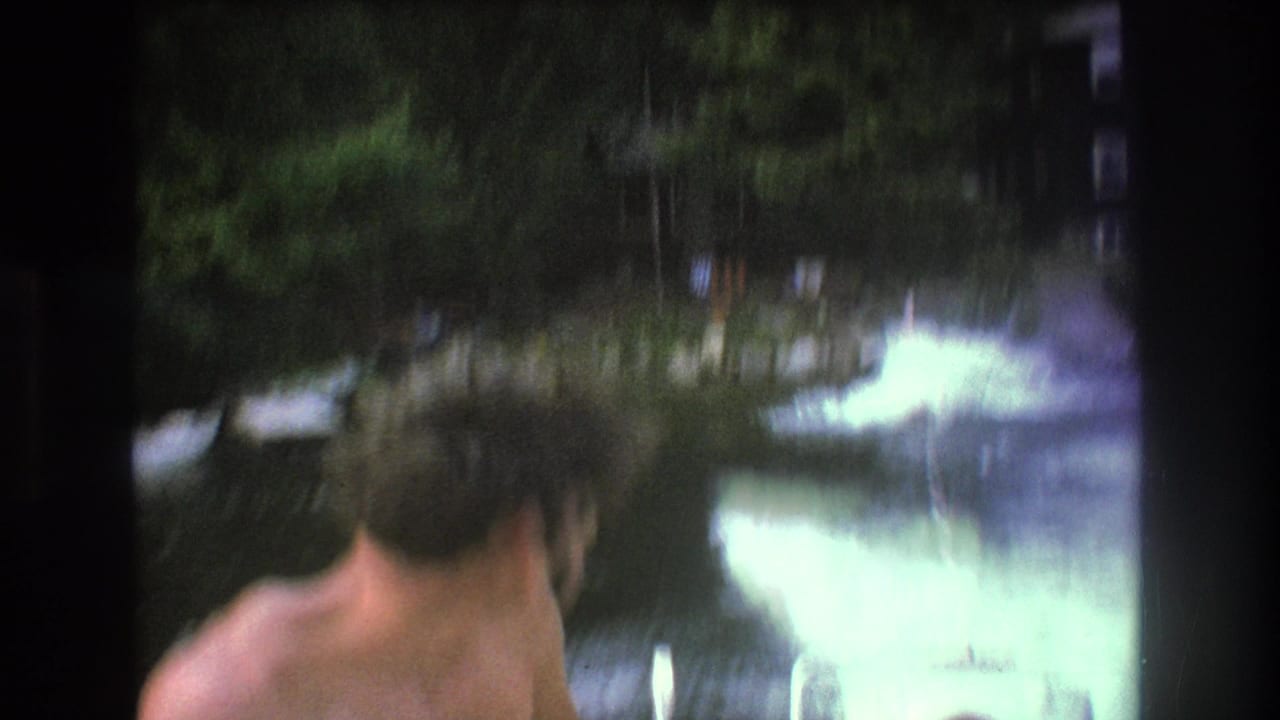 A shirtless adult man appears at the edge of a blurred 1975 8mm Boston-area lake scene, with wake, trees, and soft home-movie exposure visible.