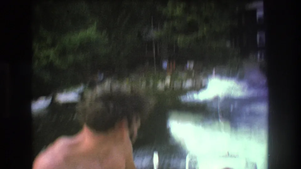 A shirtless adult man appears at the edge of a blurred 1975 8mm Boston-area lake scene, with wake, trees, and soft home-movie exposure visible.
