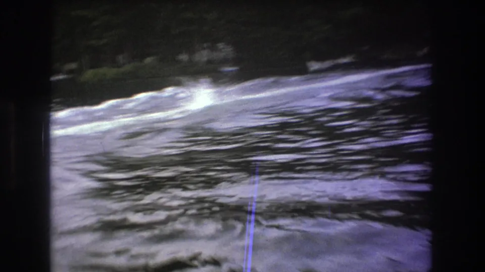 Foamy wake crosses the lake surface in a soft, grainy 1975 8mm Boston-area frame, with trees along the far shore.