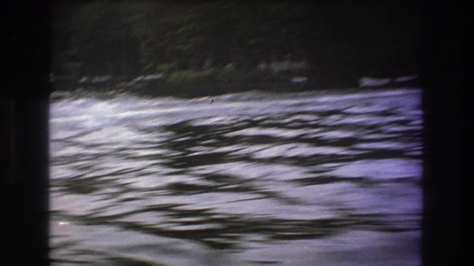 1975 8mm home-movie frame showing rippled river water and a wooded bank, with no boat clearly visible in this moment.