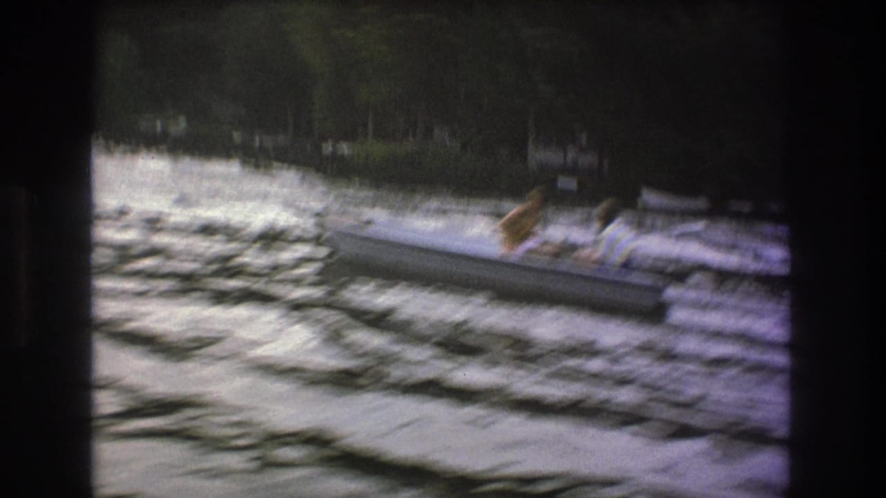 1975 8mm home-movie frame showing people in a small canoe or kayak-like boat crossing fast water near trees along the shore.