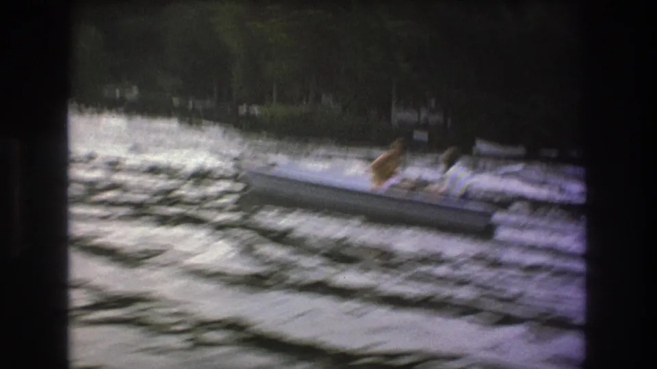 1975 8mm home-movie frame showing people in a small canoe or kayak-like boat crossing fast water near trees along the shore.