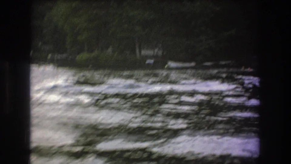 1975 8mm home-movie frame with motion-blurred whitewater and a tree-lined riverbank as the boat moves out of view.