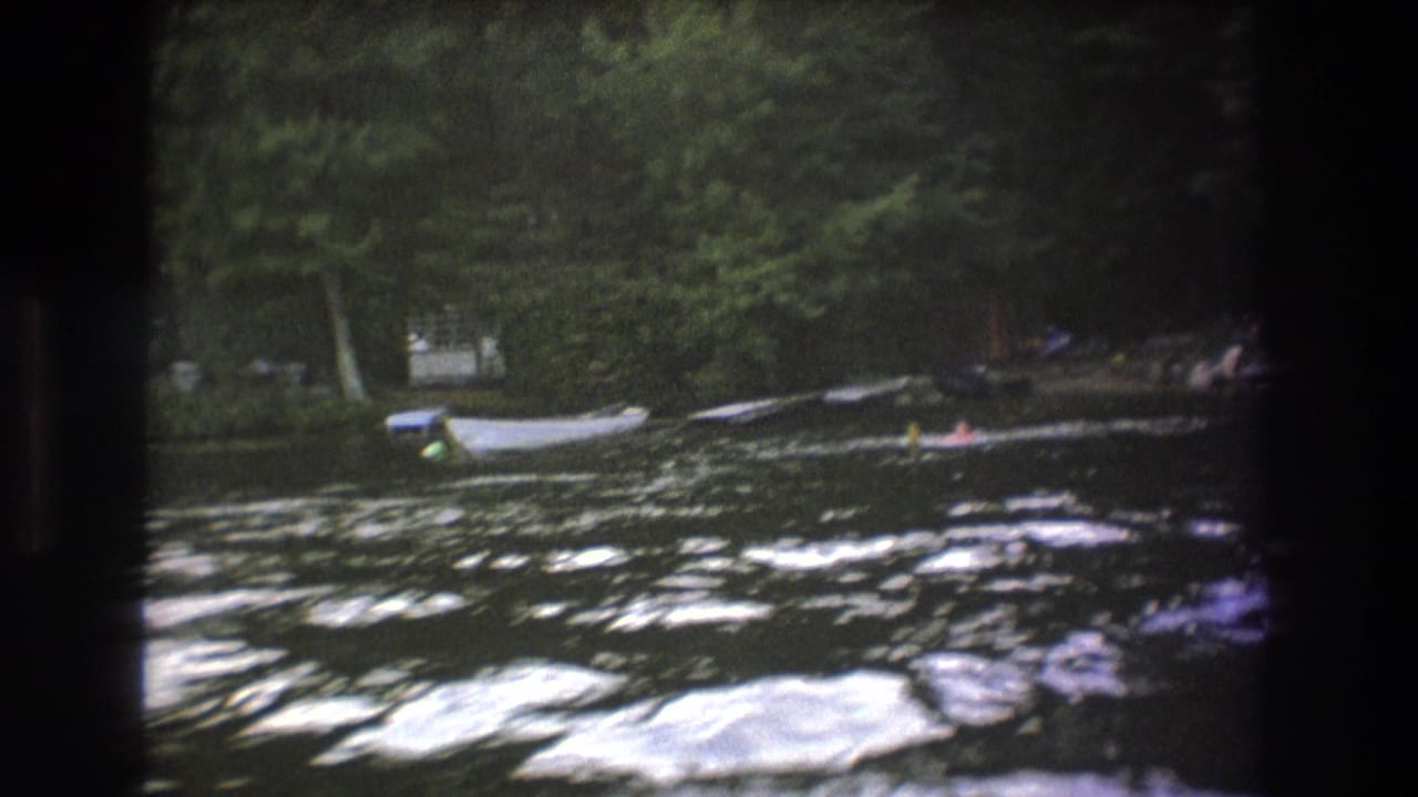 1975 Boston-area 8mm frame showing distant swimmers in rippling lake water, with small boats moored near trees on the far shore.