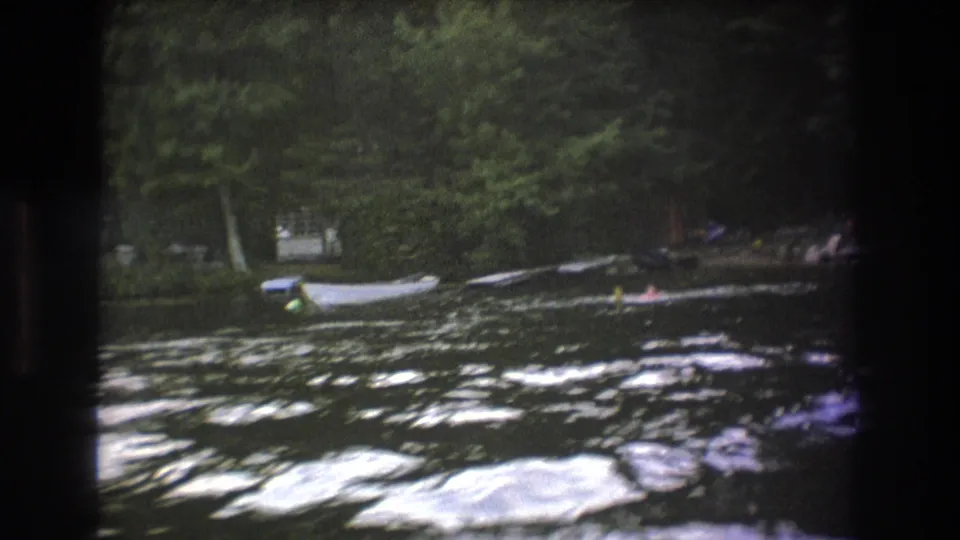 1975 Boston-area 8mm frame showing distant swimmers in rippling lake water, with small boats moored near trees on the far shore.