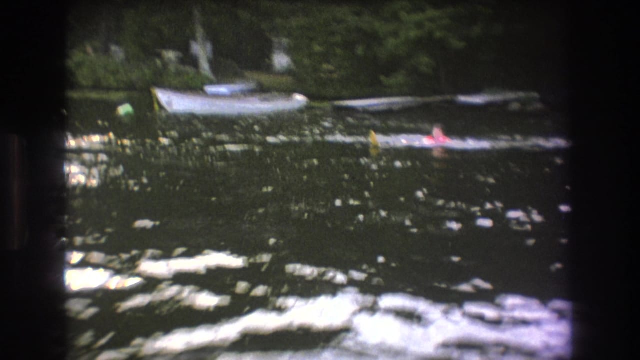 1975 Boston-area home-movie frame showing two distant swimmers in calm lake water, backed by small boats and dense green trees.