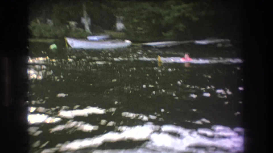 1975 Boston-area home-movie frame showing two distant swimmers in calm lake water, backed by small boats and dense green trees.