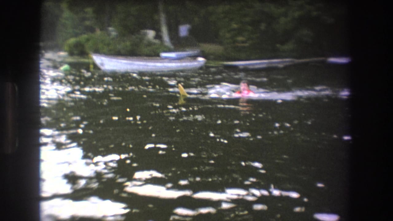 Distant swimmers move through a wooded Boston-area lake in 1975, with a white boat and darker boats moored along the far bank.