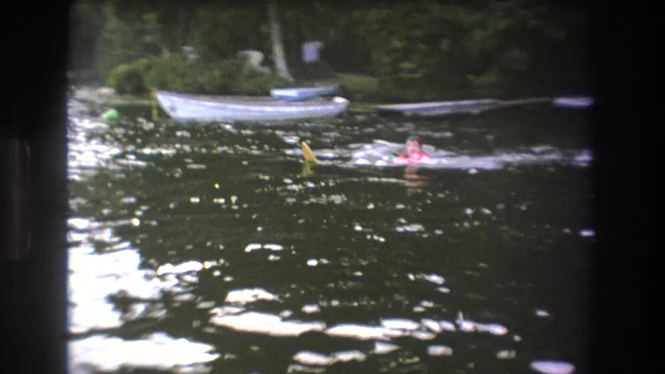 Distant swimmers move through a wooded Boston-area lake in 1975, with a white boat and darker boats moored along the far bank.