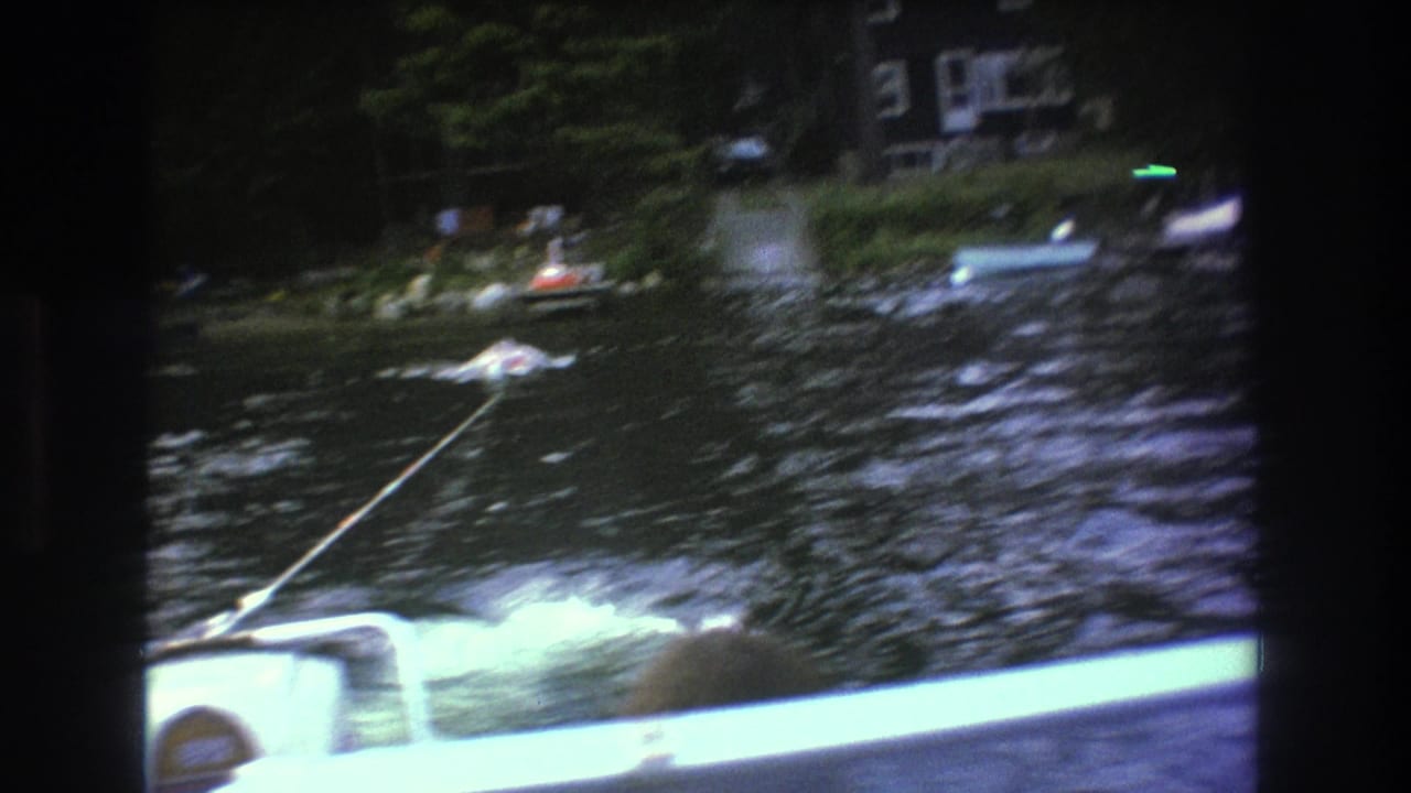 1975 8mm home-movie frame from the Boston area showing a motorboat point of view with tow rope, foamy wake, dark lake water, and distant shoreline houses among trees.