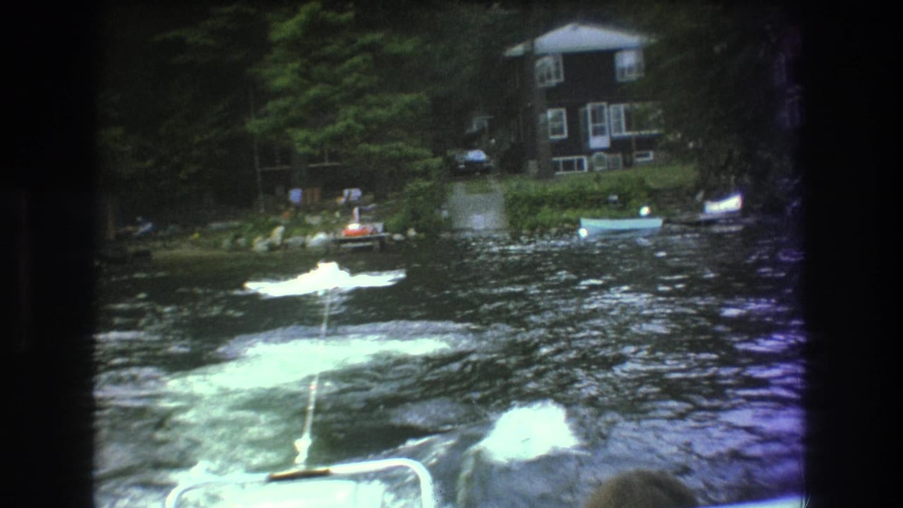 Foamy wake and tow rope behind a moving boat with lakeside houses and small boats along the shore.
