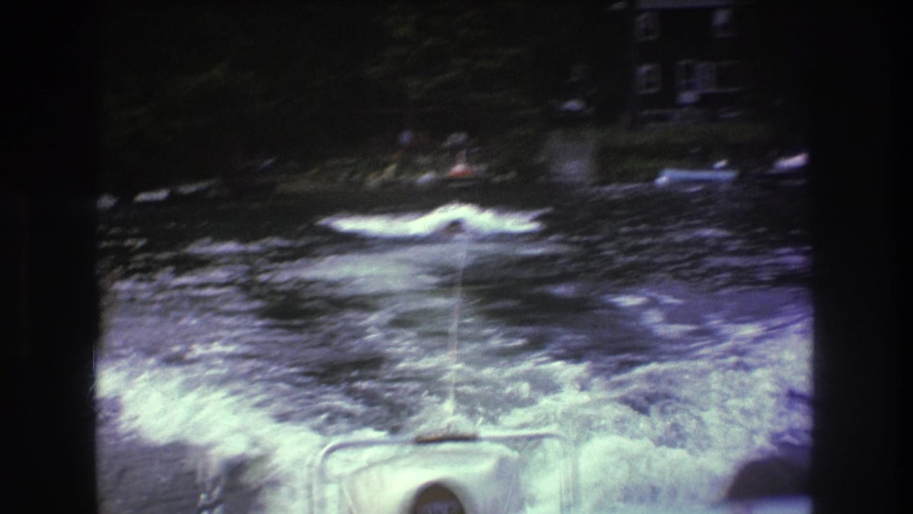 1975 8mm home-movie frame from the Boston area showing a distant person being towed behind a boat through foamy wake, with shoreline houses and trees beyond the water.