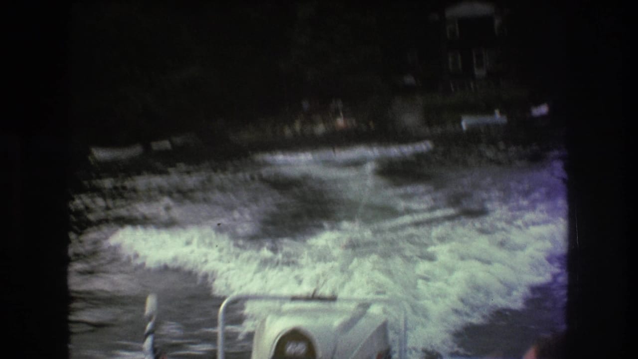 1975 Boston-area 8mm frame with strong motion blur showing white motorboat wake, a tow rope over dark lake water, and a tree-lined shore with boats and buildings in the distance.