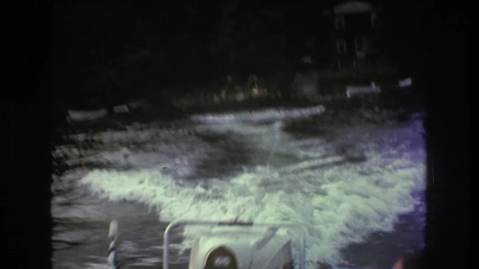 1975 Boston-area 8mm frame with strong motion blur showing white motorboat wake, a tow rope over dark lake water, and a tree-lined shore with boats and buildings in the distance.