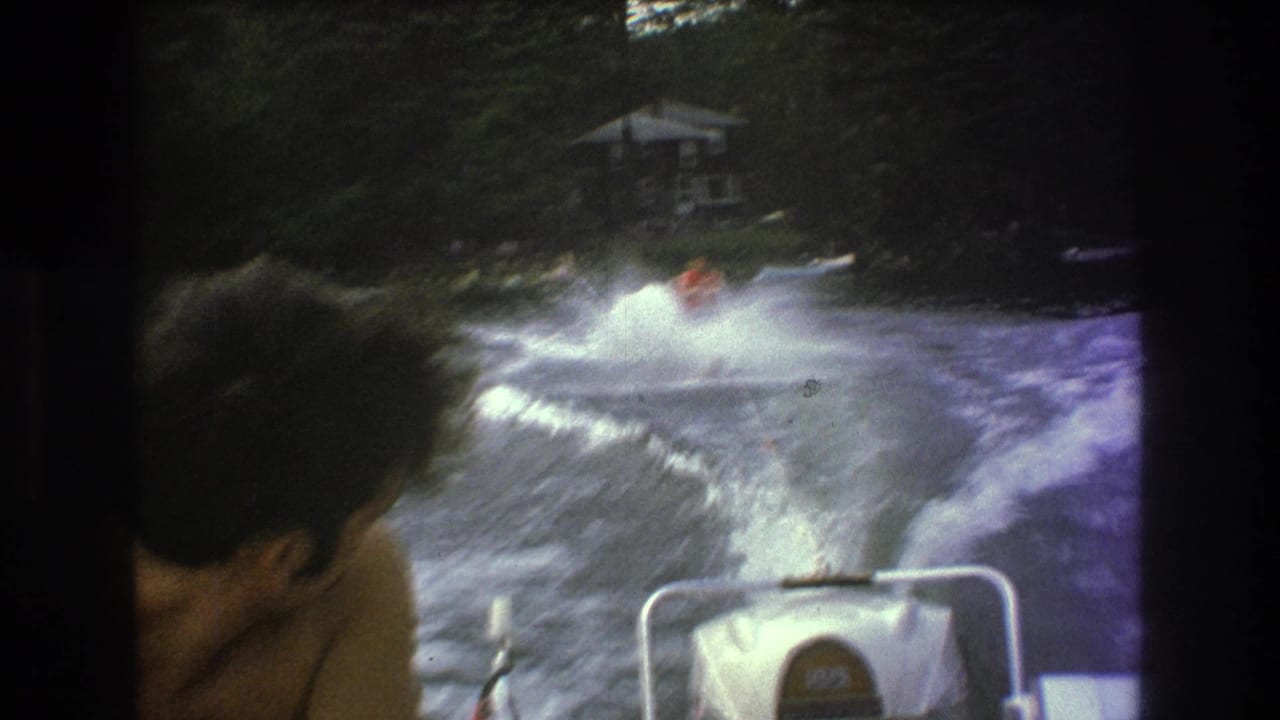 1975 8mm home-movie frame from the Boston metadata context showing a water skier behind a speedboat, with a passenger head in the foreground and wooded shoreline beyond.