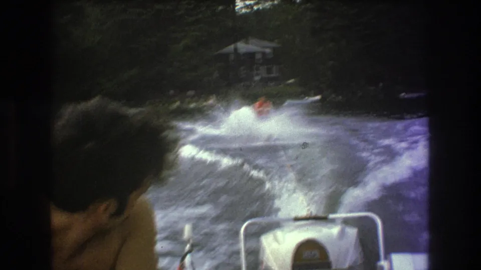 1975 8mm home-movie frame from the Boston metadata context showing a water skier behind a speedboat, with a passenger head in the foreground and wooded shoreline beyond.