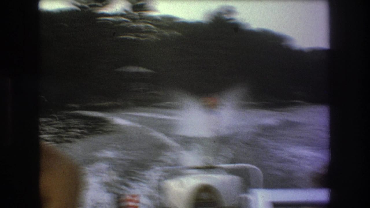 Soft, motion-blurred 8mm frame showing the skier centered in the wake behind the boat, with a foreground passenger edge, stern rail, and wooded shoreline in view.