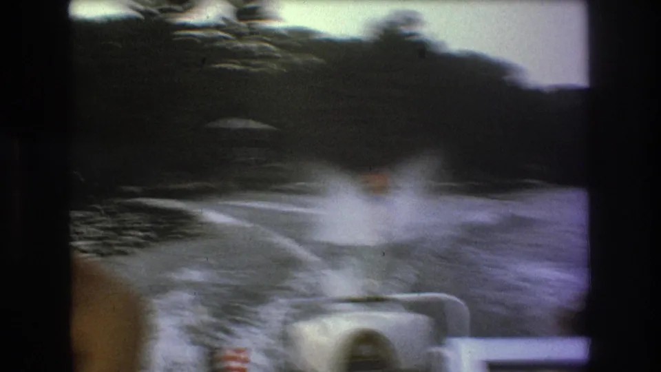 Soft, motion-blurred 8mm frame showing the skier centered in the wake behind the boat, with a foreground passenger edge, stern rail, and wooded shoreline in view.