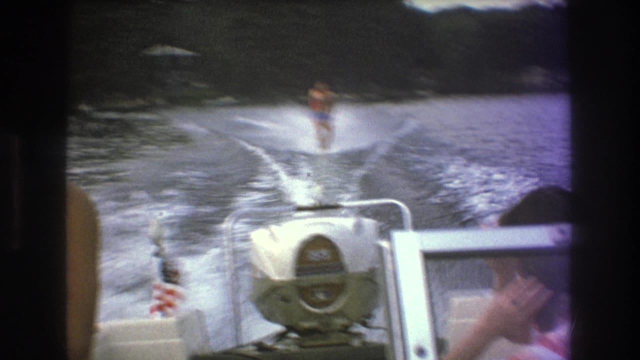 Boston metadata-context 1975 home-movie frame showing a skier in the wake behind a speedboat, with a stern emblem, American flag, seated passenger, and wooded shoreline visible.