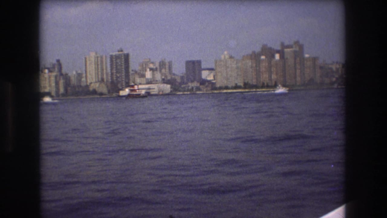 Boston waterfront skyline seen across open water in 1975 8mm footage, with a larger vessel near the buildings and dark film borders visible.