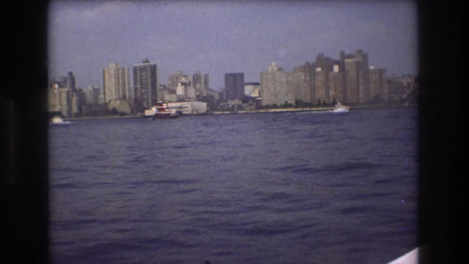Boston waterfront skyline seen across open water in 1975 8mm footage, with a larger vessel near the buildings and dark film borders visible.