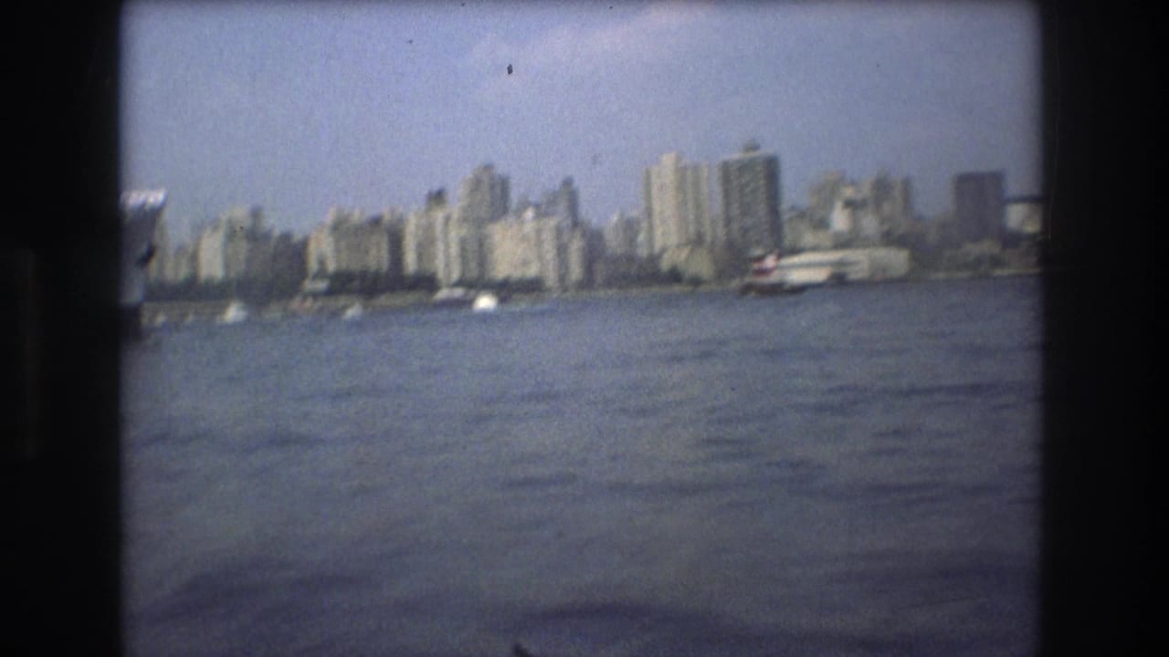 Boston waterfront skyline in 1975 seen from across the water, with high-rise buildings, small boats, hazy sky, and rounded dark 8mm frame edges.