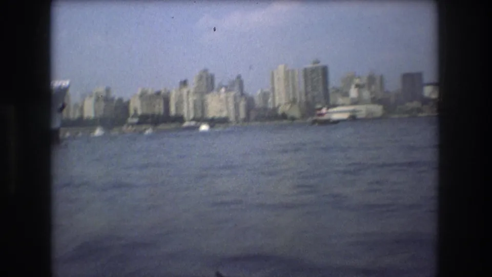 Boston waterfront skyline in 1975 seen from across the water, with high-rise buildings, small boats, hazy sky, and rounded dark 8mm frame edges.