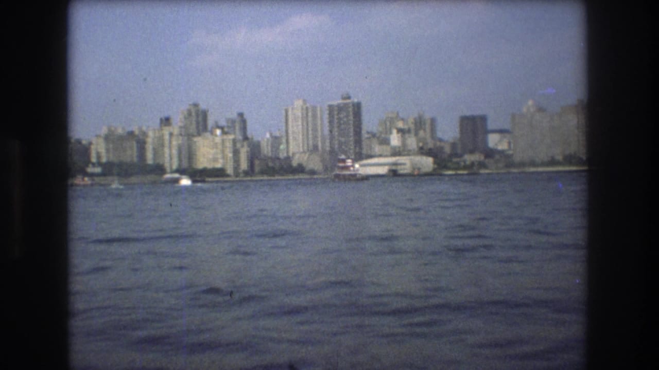 1975 Boston skyline viewed from a moving boat, with rippled harbor water, dense waterfront buildings, and a red-and-white ferry-like vessel near shore.