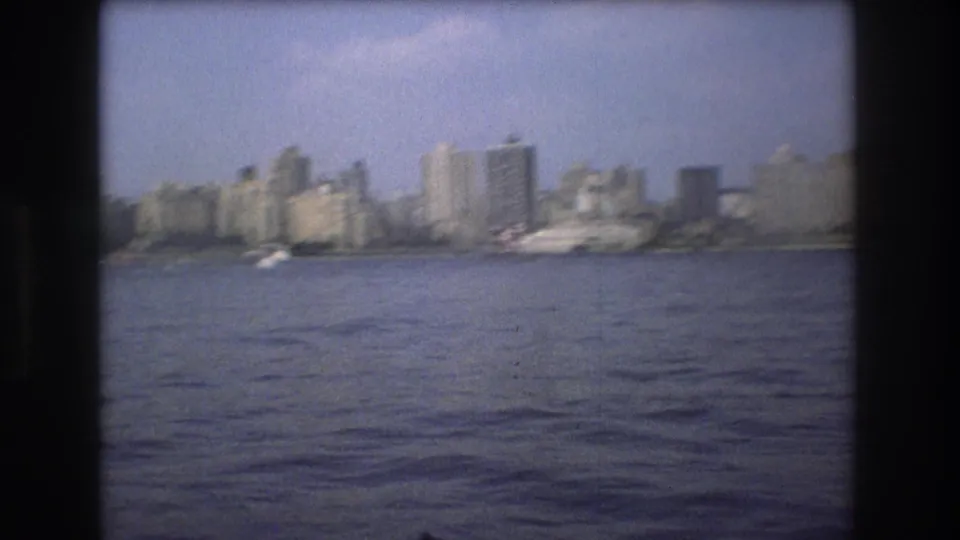 Soft 8mm view of Boston in 1975 from the harbor, showing a distant skyline, small boats, and broad water in the foreground.