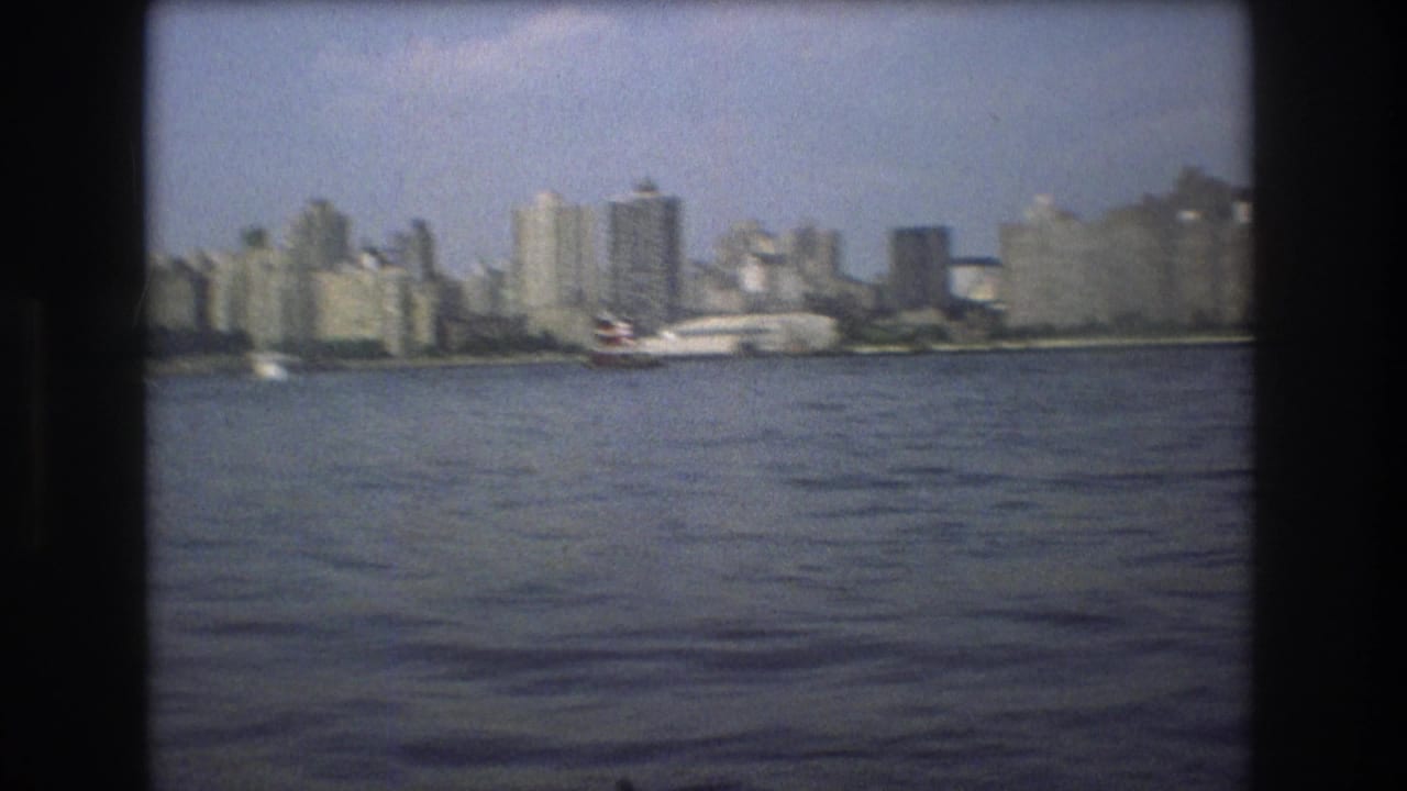 Boston waterfront skyline in 1975 with rippled harbor water, clustered high-rises, a ferry-like vessel near shore, and visible 8mm film softness.