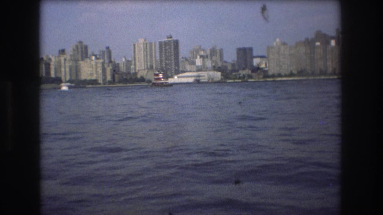 1975 Boston home-movie frame showing rippled harbor water, a red-and-white vessel, and a distant waterfront skyline under hazy daylight.
