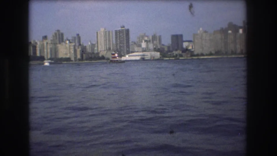 1975 Boston home-movie frame showing rippled harbor water, a red-and-white vessel, and a distant waterfront skyline under hazy daylight.