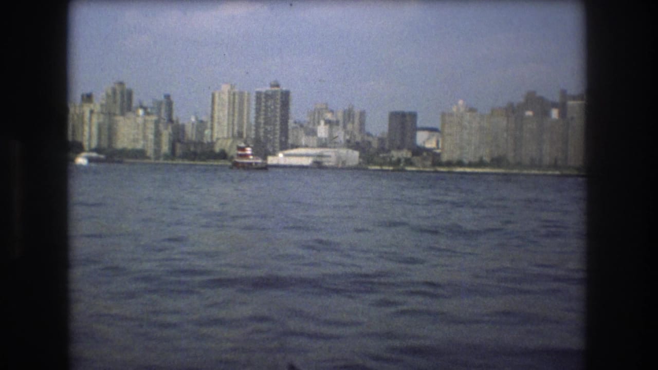 1975 Boston waterfront view across open harbor water, with a small red-and-white boat and rows of tall buildings along the shore.
