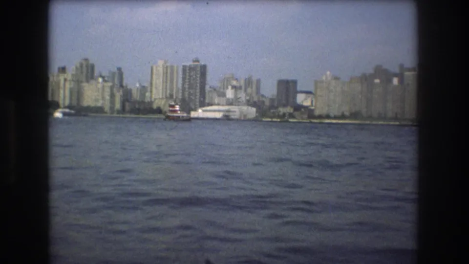 1975 Boston waterfront view across open harbor water, with a small red-and-white boat and rows of tall buildings along the shore.