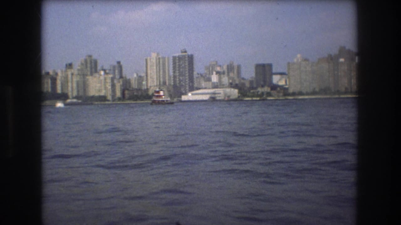 1975 Boston harbor frame with rippled water in the foreground, a red-and-white vessel near shore, and high-rise buildings across the skyline.