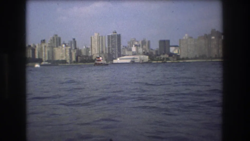 1975 Boston harbor frame with rippled water in the foreground, a red-and-white vessel near shore, and high-rise buildings across the skyline.