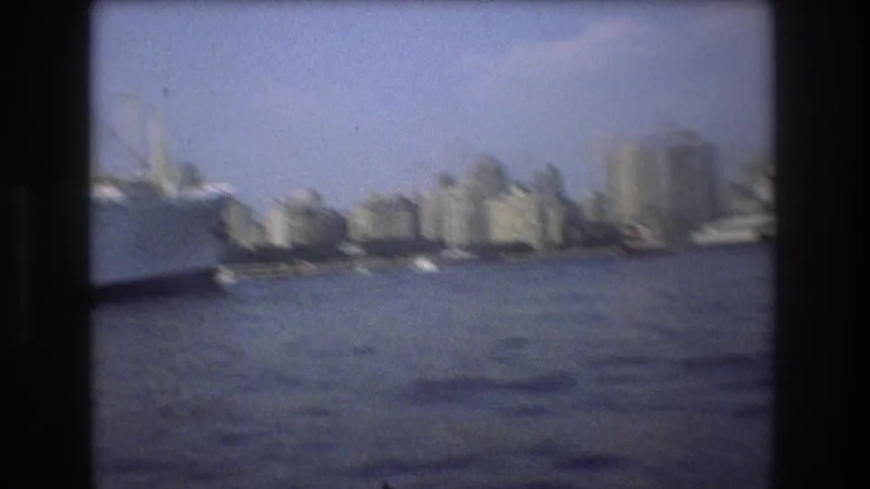 Soft 8mm frame of Boston harbor in 1975 showing the city skyline across the water and a large vessel partly entering from the left edge.