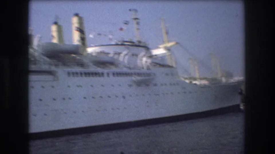Large white ship in Boston harbor in 1975, seen close to frame with smokestacks, deck structures, and harbor cranes in the background.