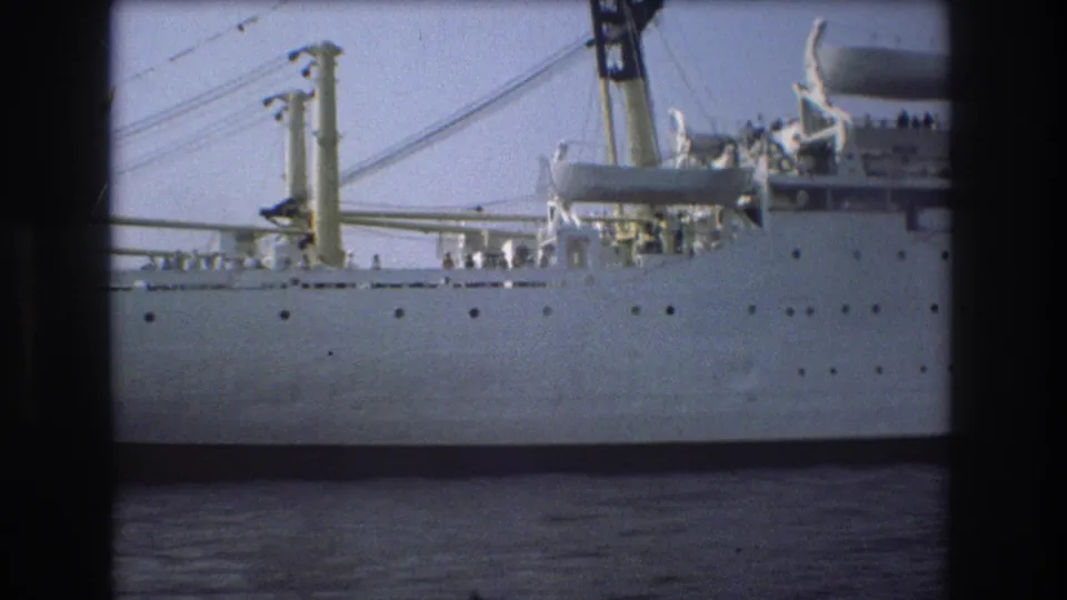 1975 Boston 8mm frame showing the side of a large white ship on harbor water, with rows of portholes, davits, deck gear, and small figures visible near the rail.