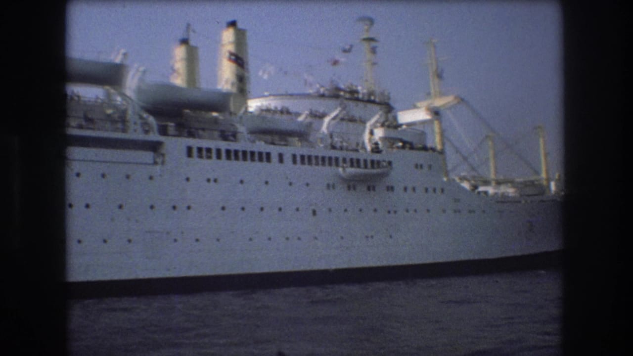 1975 Boston 8mm frame showing the large white ship continuing past the camera, with funnels, lifeboat gear, rigging, portholes, and harbor water visible.
