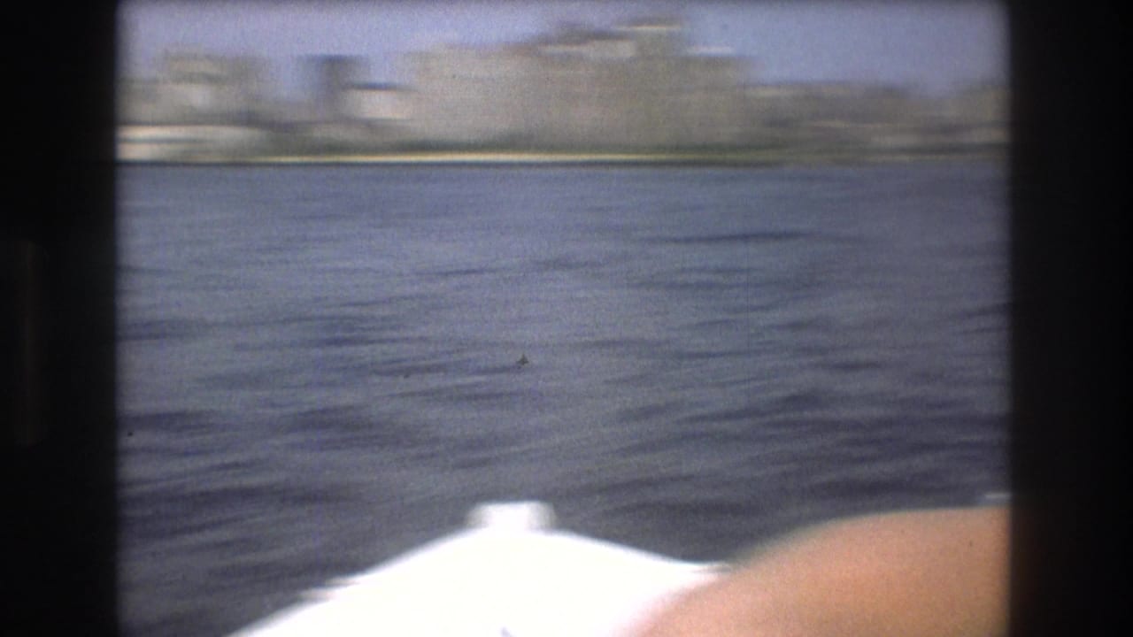 A handheld pan or motion-blurred view shows harbor water, a distant Boston waterfront, and a bright foreground edge of the filming boat.
