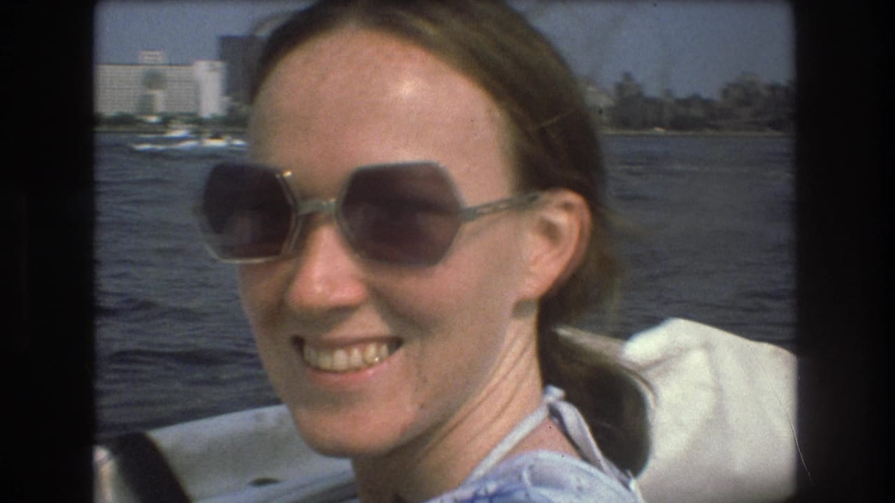 A smiling adult woman wearing large sunglasses sits on a boat in Boston harbor, with water and distant waterfront buildings behind her in the 1975 home-movie footage.